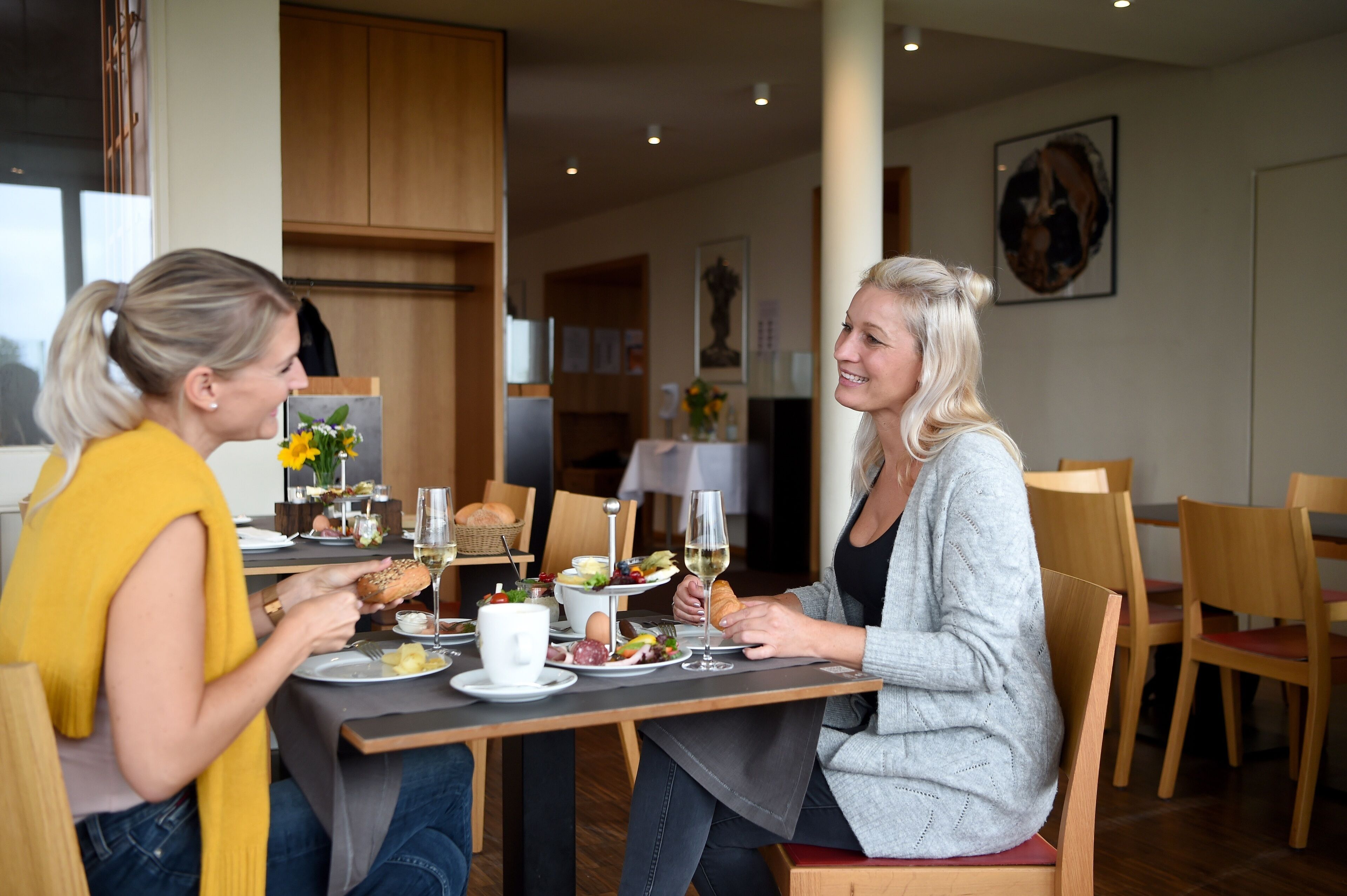 Zwei Personen essen an einem Tisch mit verschiedenen Speisen und Getränken, in einem Raum mit Holzmöbeln und einem Bild an der Wand.