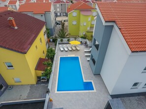 A residential area with a swimming pool surrounded by colorful houses, a small patio with lounge chairs, and a yellow building with a chimney.