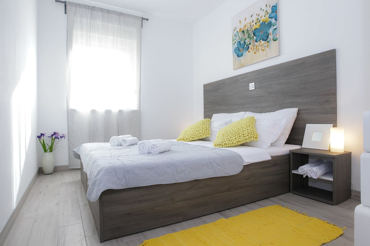 A modern bedroom with a wooden headboard, a bed with white bedding and yellow pillows, a bedside table with a lamp, and a yellow rug.