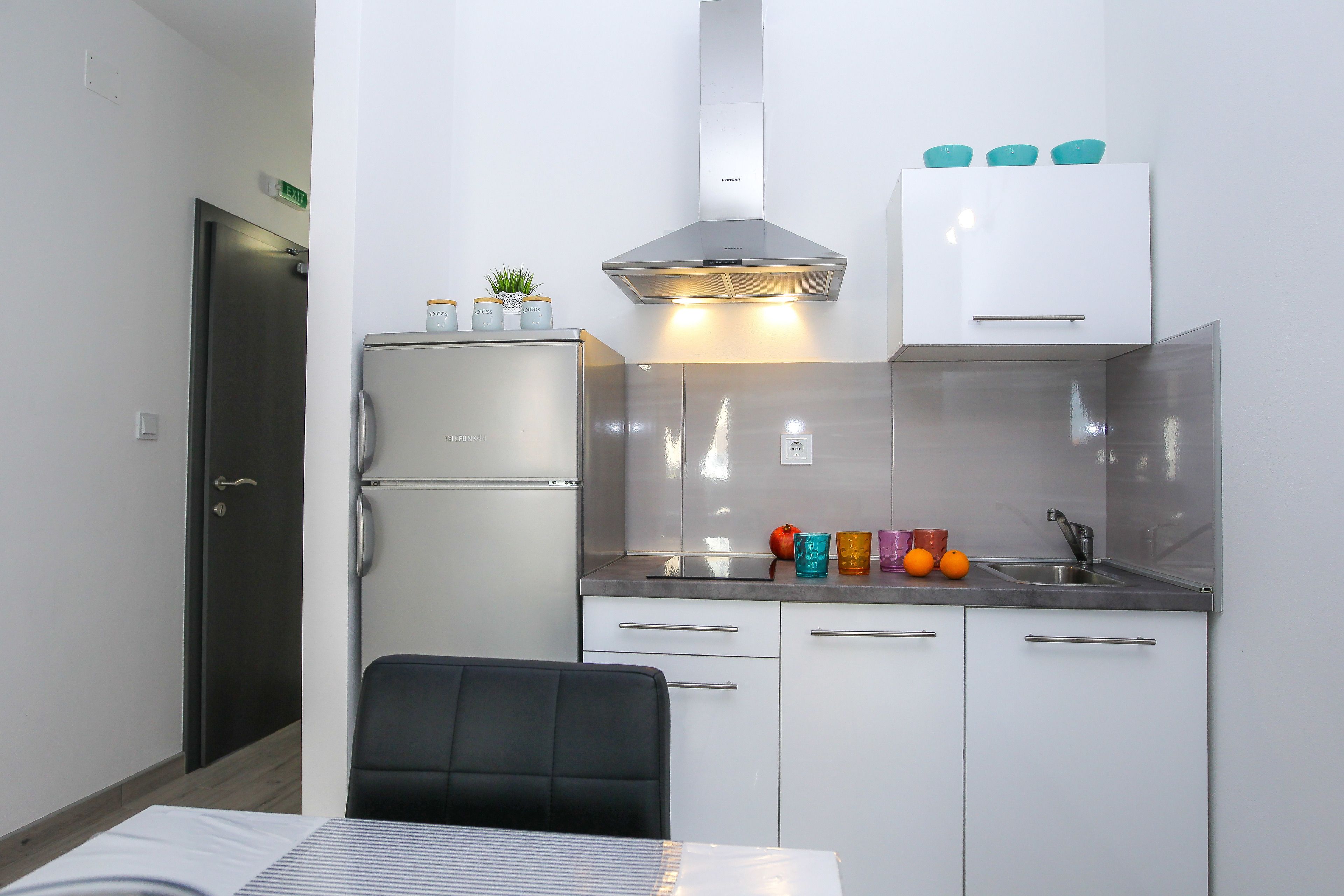 A compact kitchenette with a stainless steel refrigerator, a built-in oven, a range hood, and a countertop with a sink and colorful glassware.