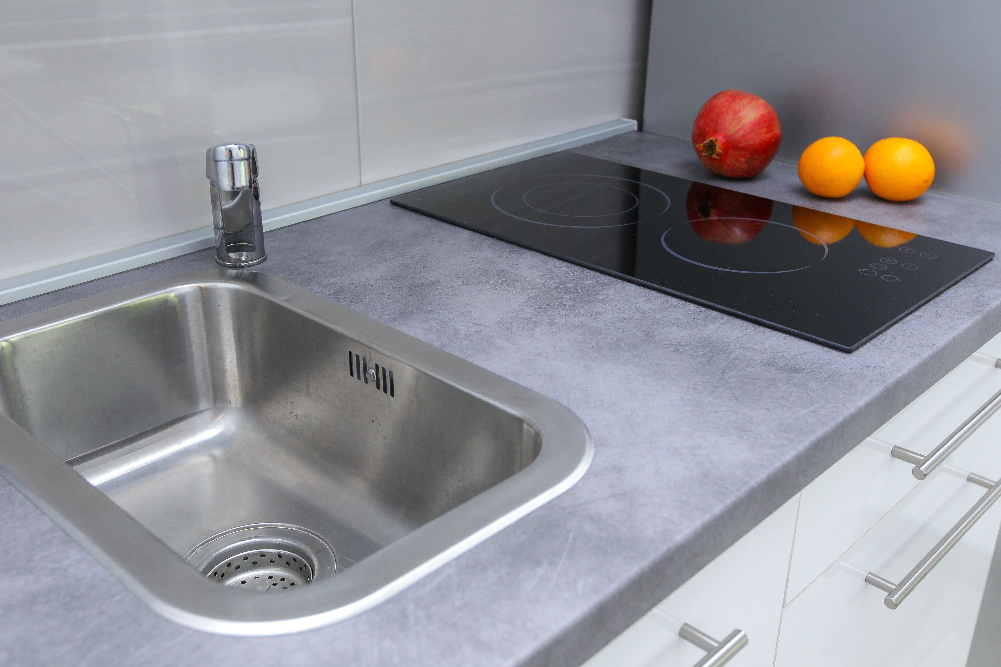 Een moderne keuken met een roestvrijstalen spoelbak, een zwart inductiekookplaatje en een granaatappel en twee sinaasappels op het aanrecht.
