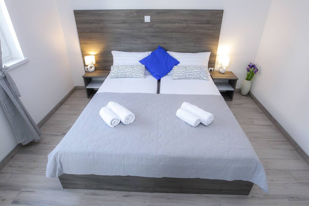 A double bed with a grey bedspread, two white pillows, and a blue accent pillow. There are two bedside tables with lamps, a vase with flowers, and towels on the bed.