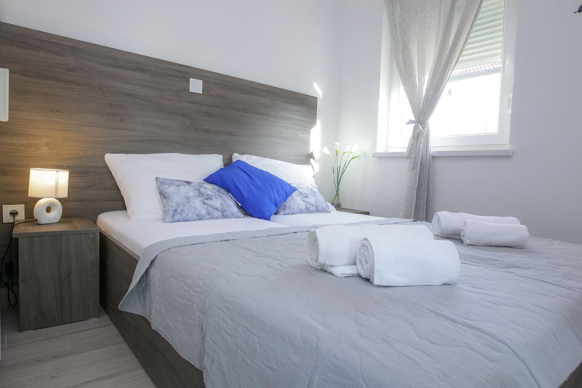 A neatly made bed with white and blue pillows, a grey bedspread, and rolled white towels on the side.