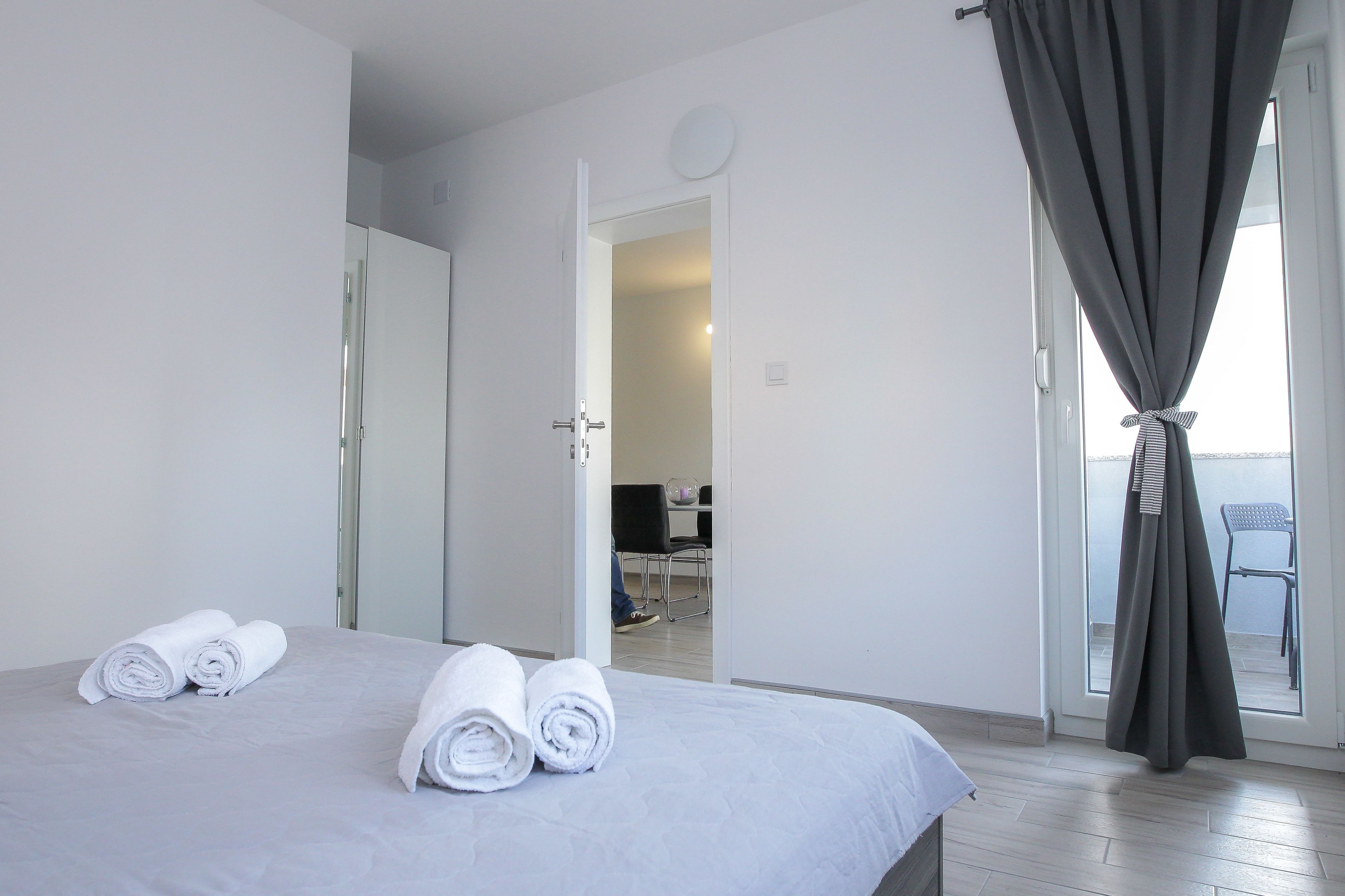Ein Hotelzimmer mit einem Bett, zwei gerollten Handtüchern, einer Tür, die auf einen Balkon führt, und einem Stuhl davor.