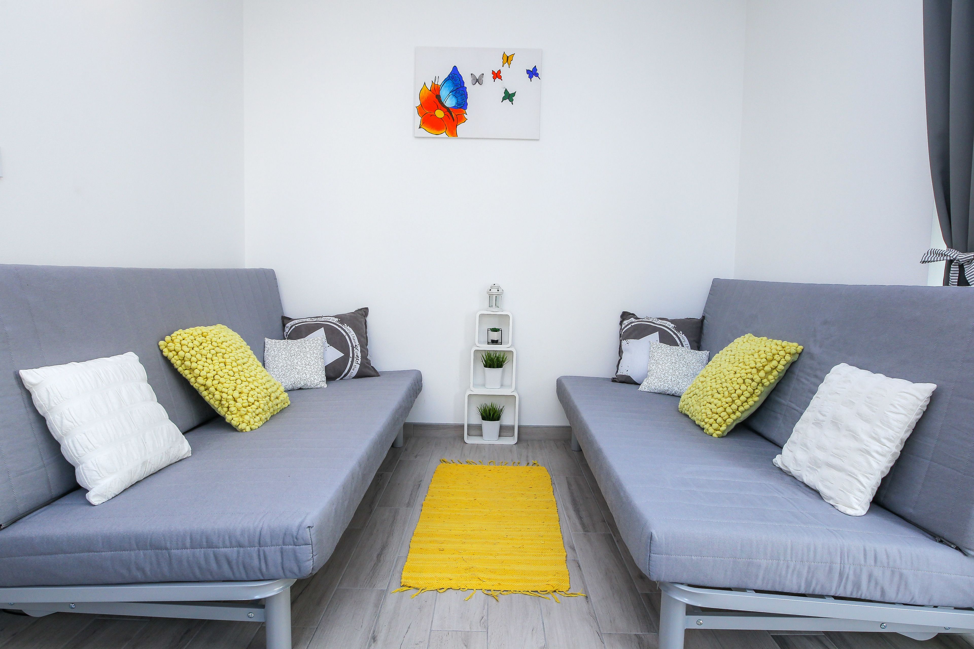 Una sala moderna con dos sofás grises, un tapete amarillo y un estante blanco pequeño con una planta.