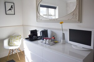 A room with a white desk, a white chair, a framed mirror, a window with a blue blind, and a flat-screen TV.