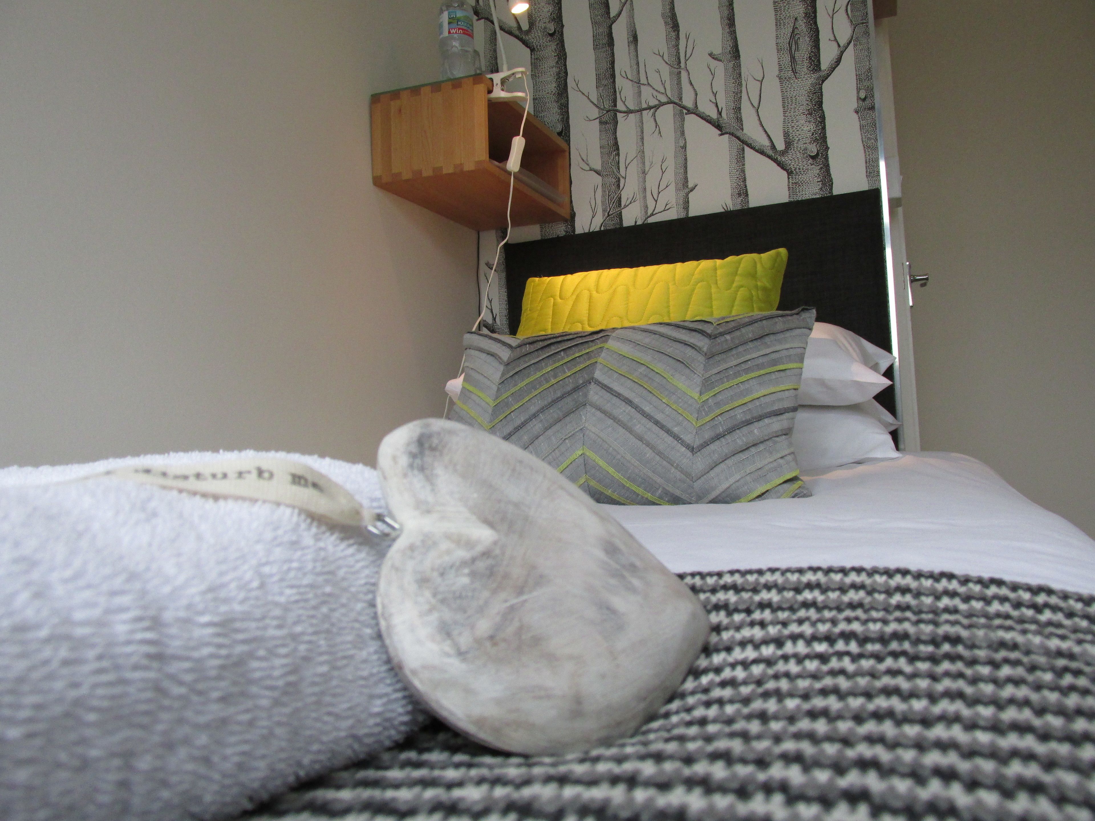 A neatly made bed with a gray and white striped blanket, a gray pillow, and a yellow pillow. A wooden shelf with a bottle and a white charger is mounted on the wall above the bed.