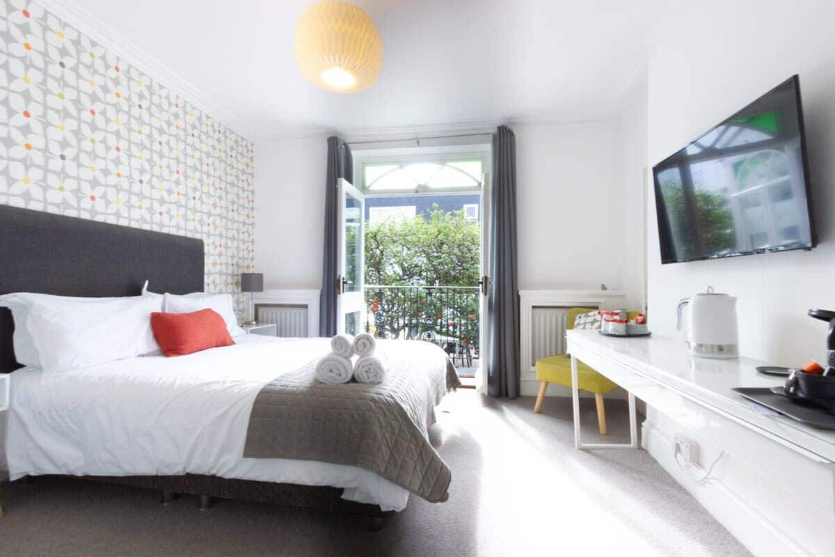 A hotel room with a bed, a desk, a TV, and a balcony with a view of trees.