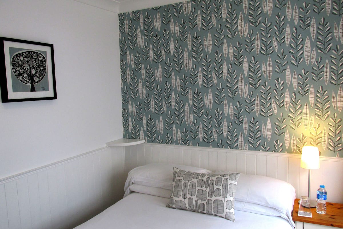 A neatly made bed with a pillow, a bedside table with a lamp and a water bottle, and a wall with a leaf pattern.