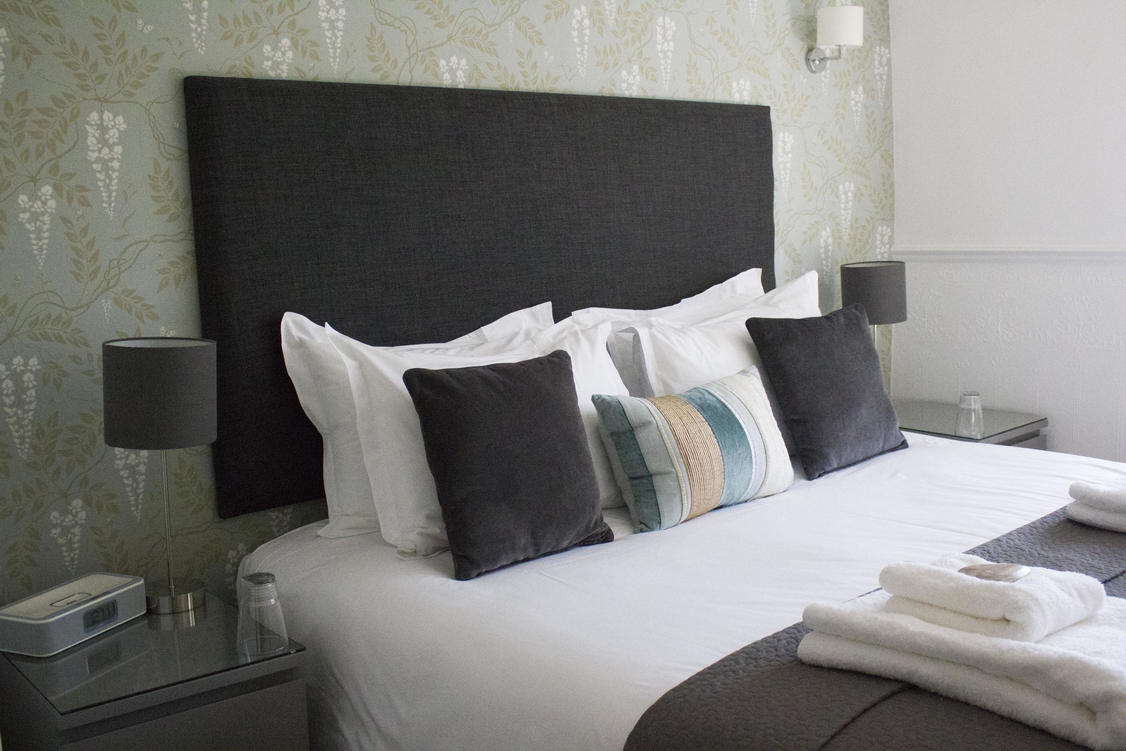 A neatly made bed with white linens and multiple pillows, a bedside table with a lamp, and a nightstand with a clock and a glass.