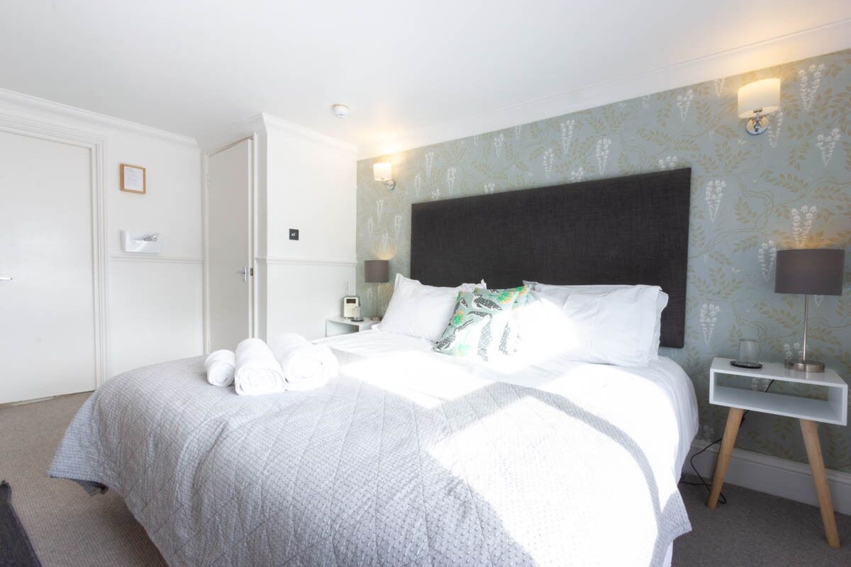 A hotel room with a large bed, white walls, a patterned wallpaper, a bedside table with a lamp, and a door.