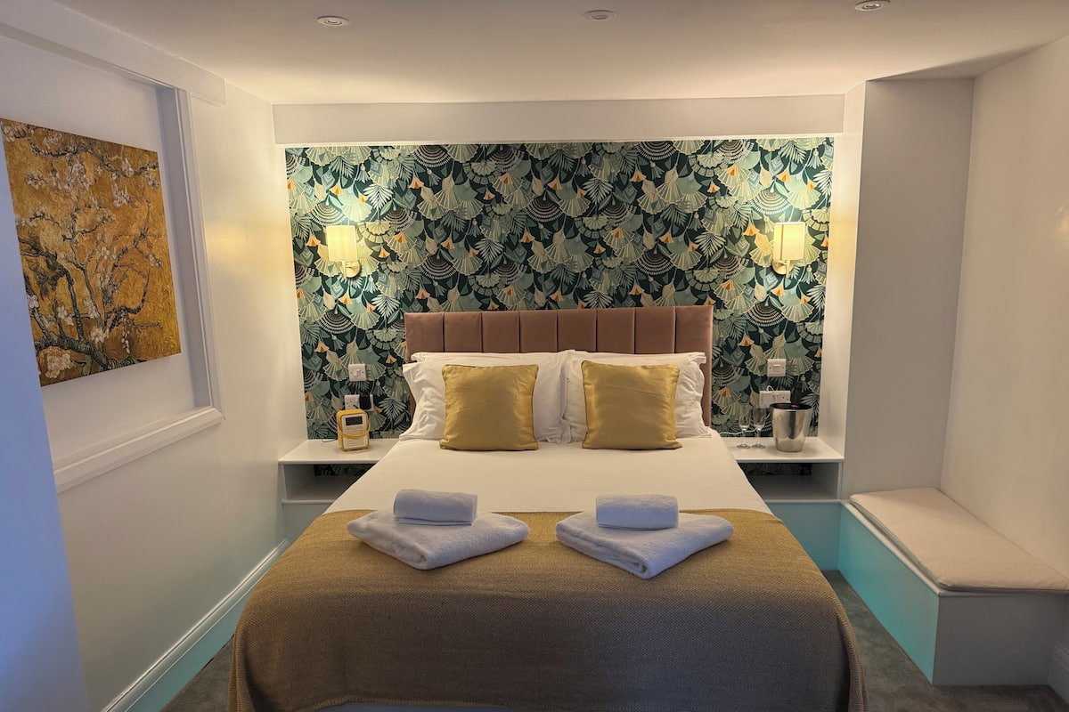 A hotel room with a bed, two bedside tables, a wall with a leafy pattern, and a painting on the wall.