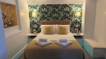 A hotel room with a bed, two bedside tables, a wall with a leafy pattern, and a painting on the wall.