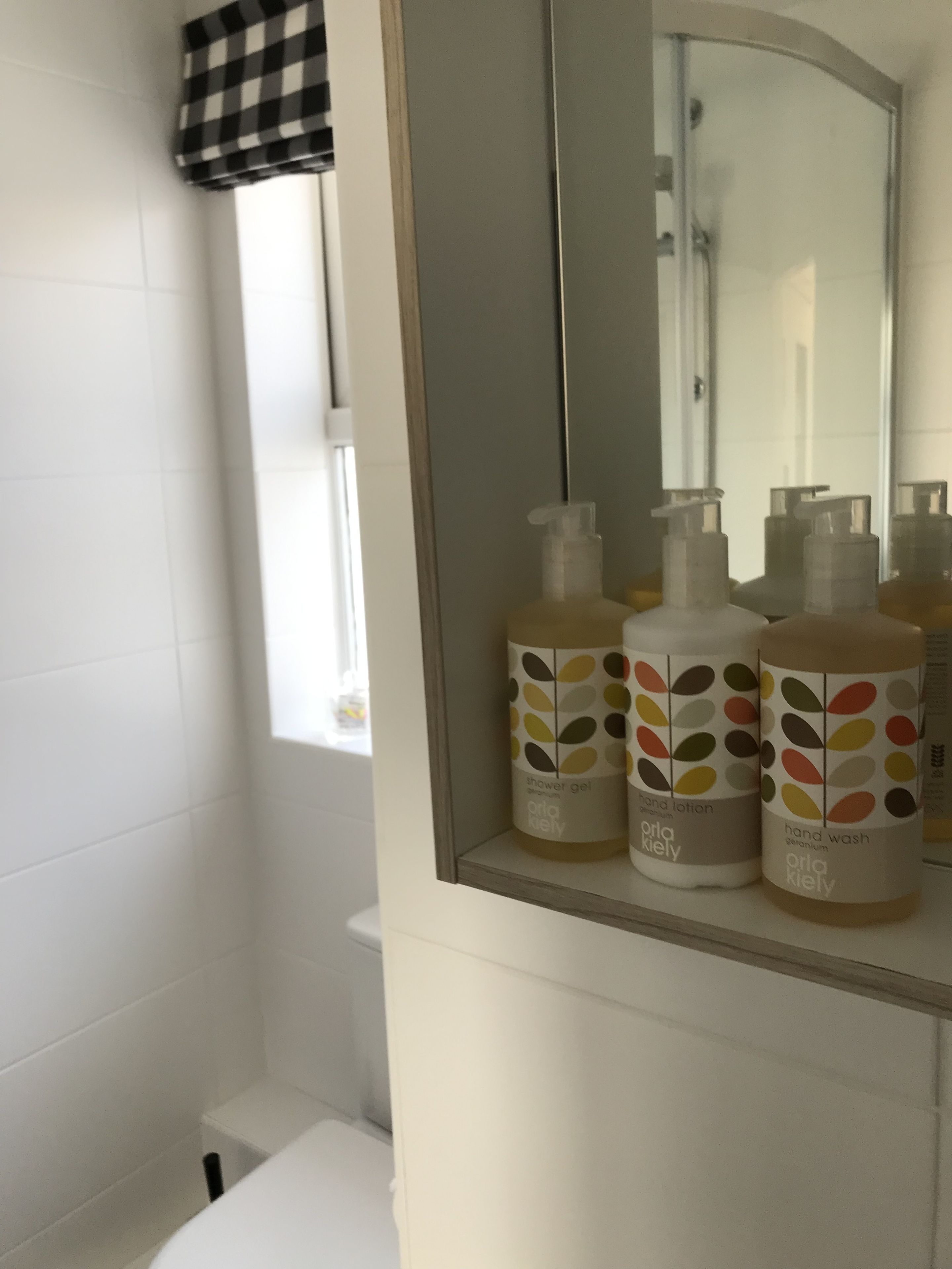 Un baño con azulejos blancos, un espejo y tres botellas de jabón de manos y gel de ducha Orla Kiley sobre una repisa.