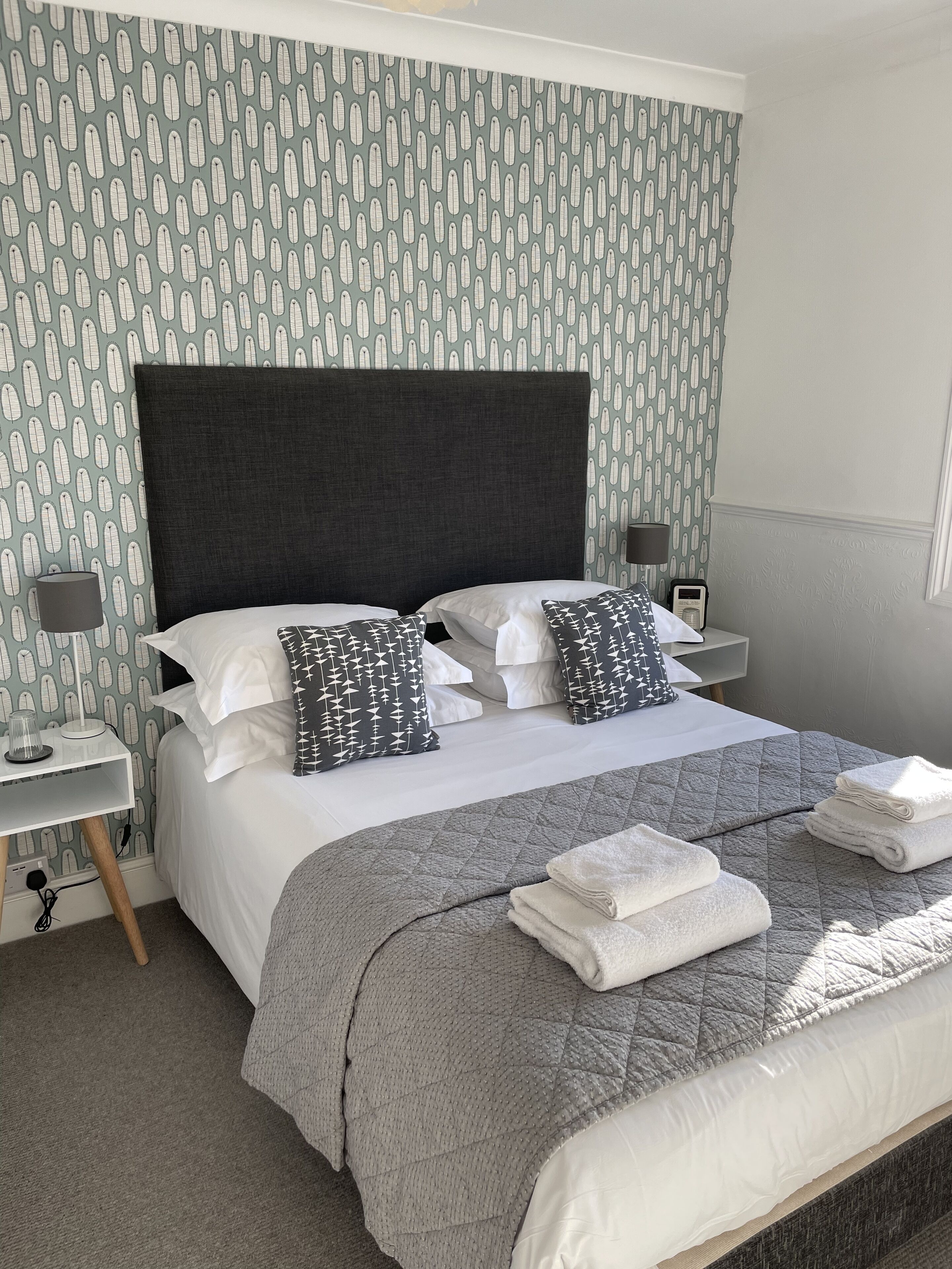 A neatly made bed with a dark headboard, white and patterned pillows, and a grey quilt. There are two bedside tables with lamps and a small speaker. The room has patterned wallpaper and a carpeted floor.
