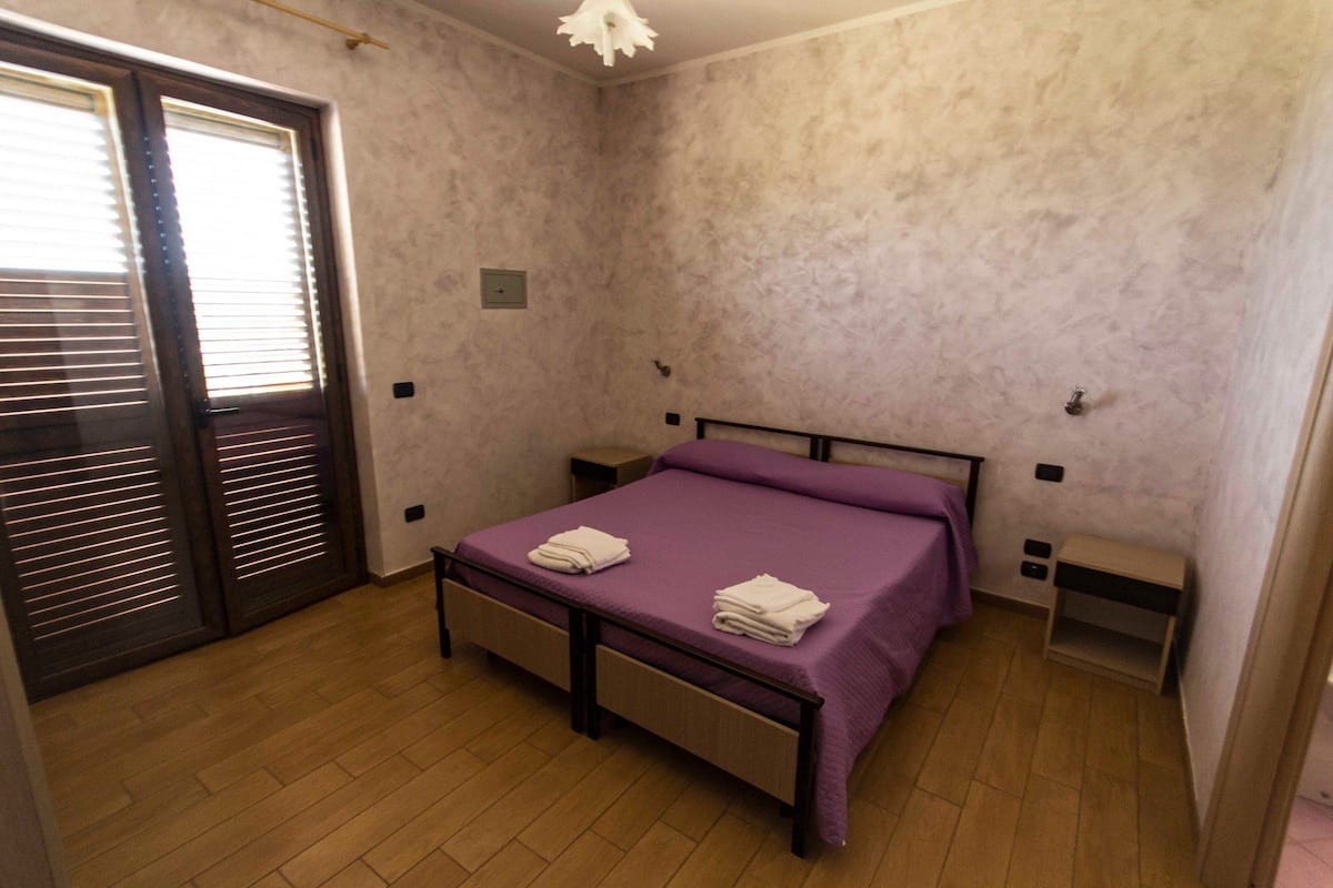 Un dormitorio con cama, mesita de noche, piso de madera y una puerta con persianas.