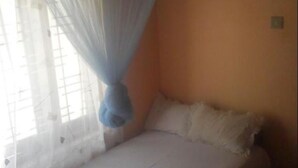 A single bed with a white mattress and pillows, a mosquito net hanging above, and a window with bars. - Regina Holiday Home (Thissamaharama)