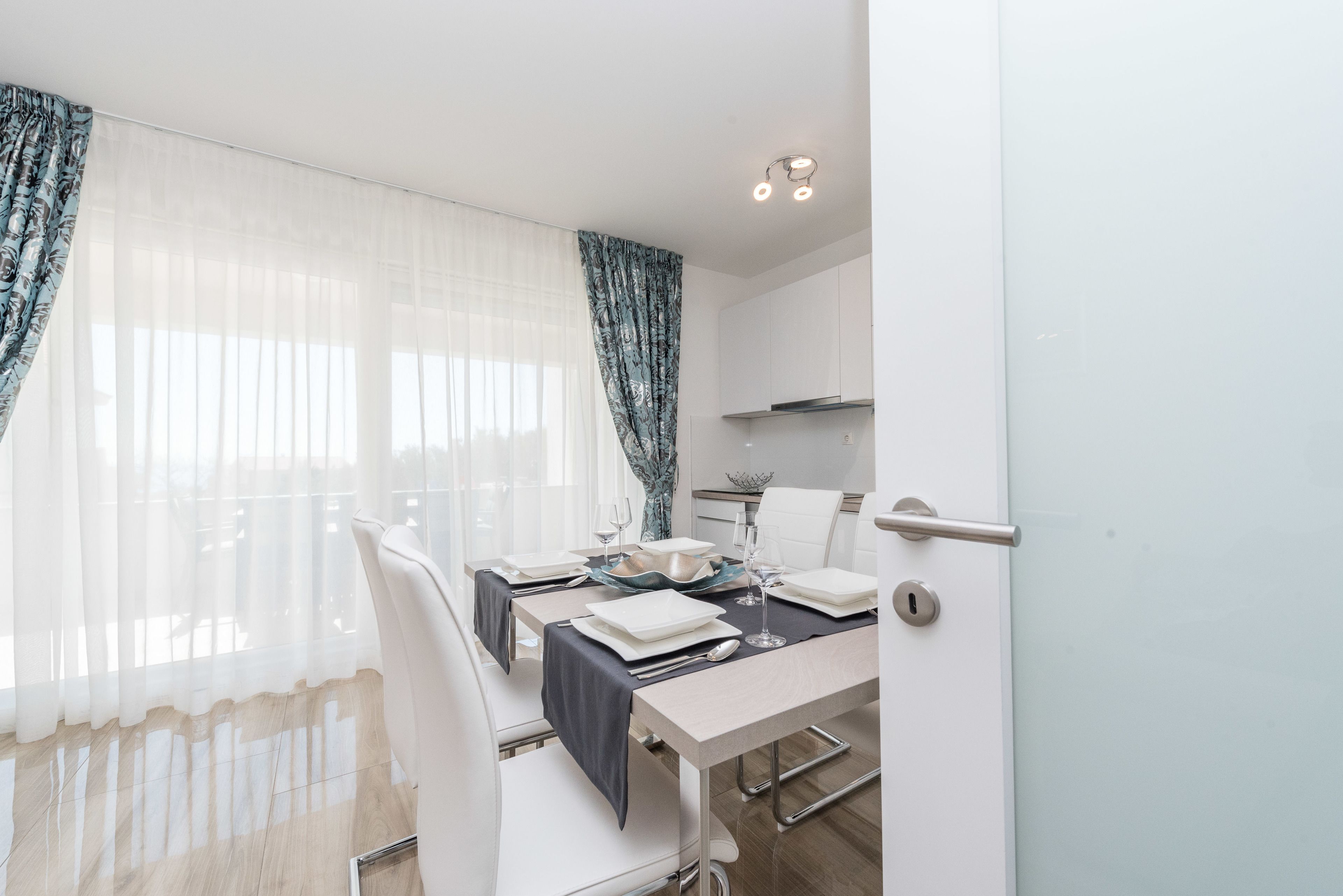 A modern dining area with a table set for two, white chairs, and a view through sheer curtains.