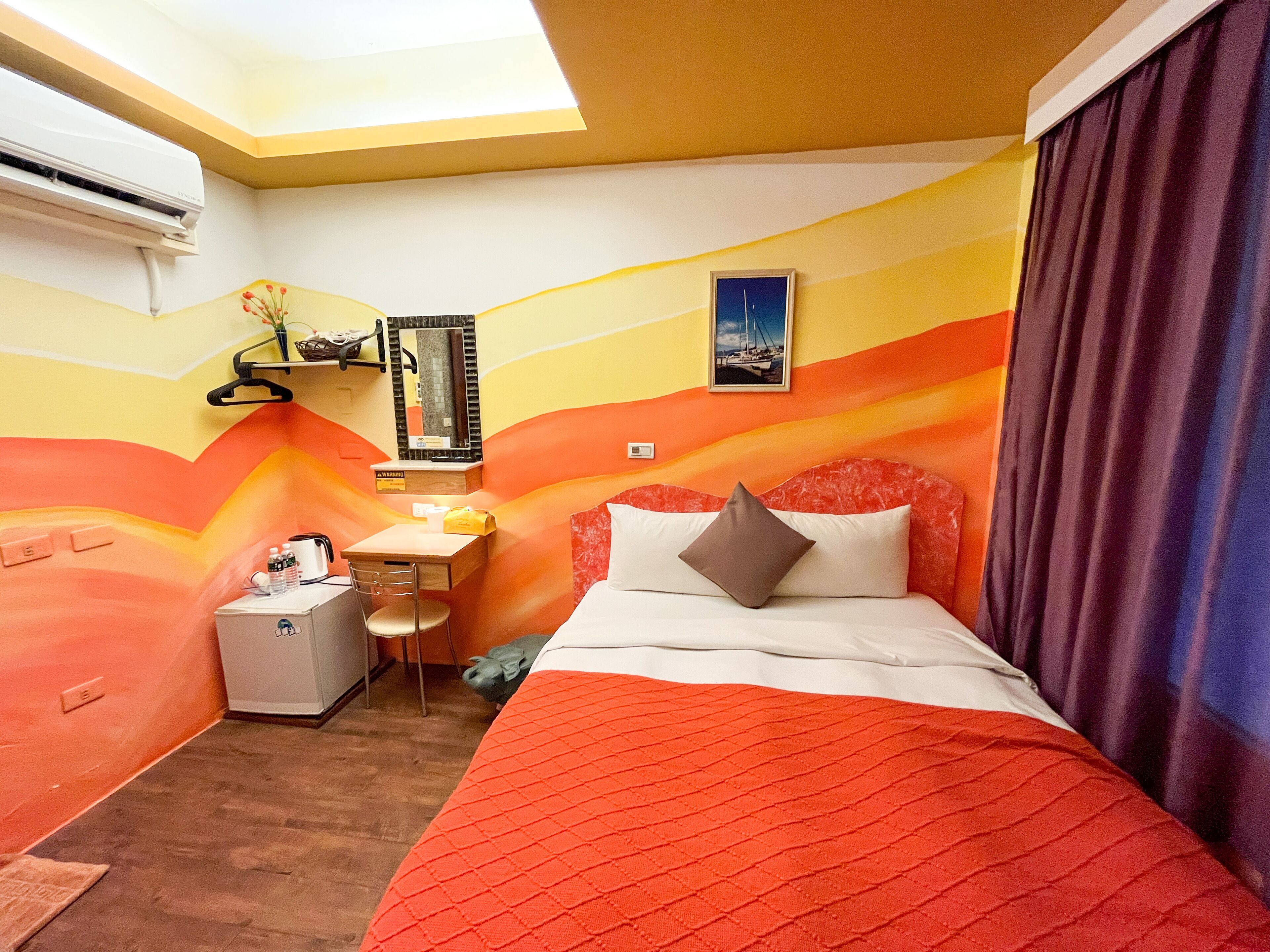 Habitación de hotel con cama, escritorio y silla. Se ve una ventana con cortinas, un aire acondicionado de pared y un cuadro enmarcado.