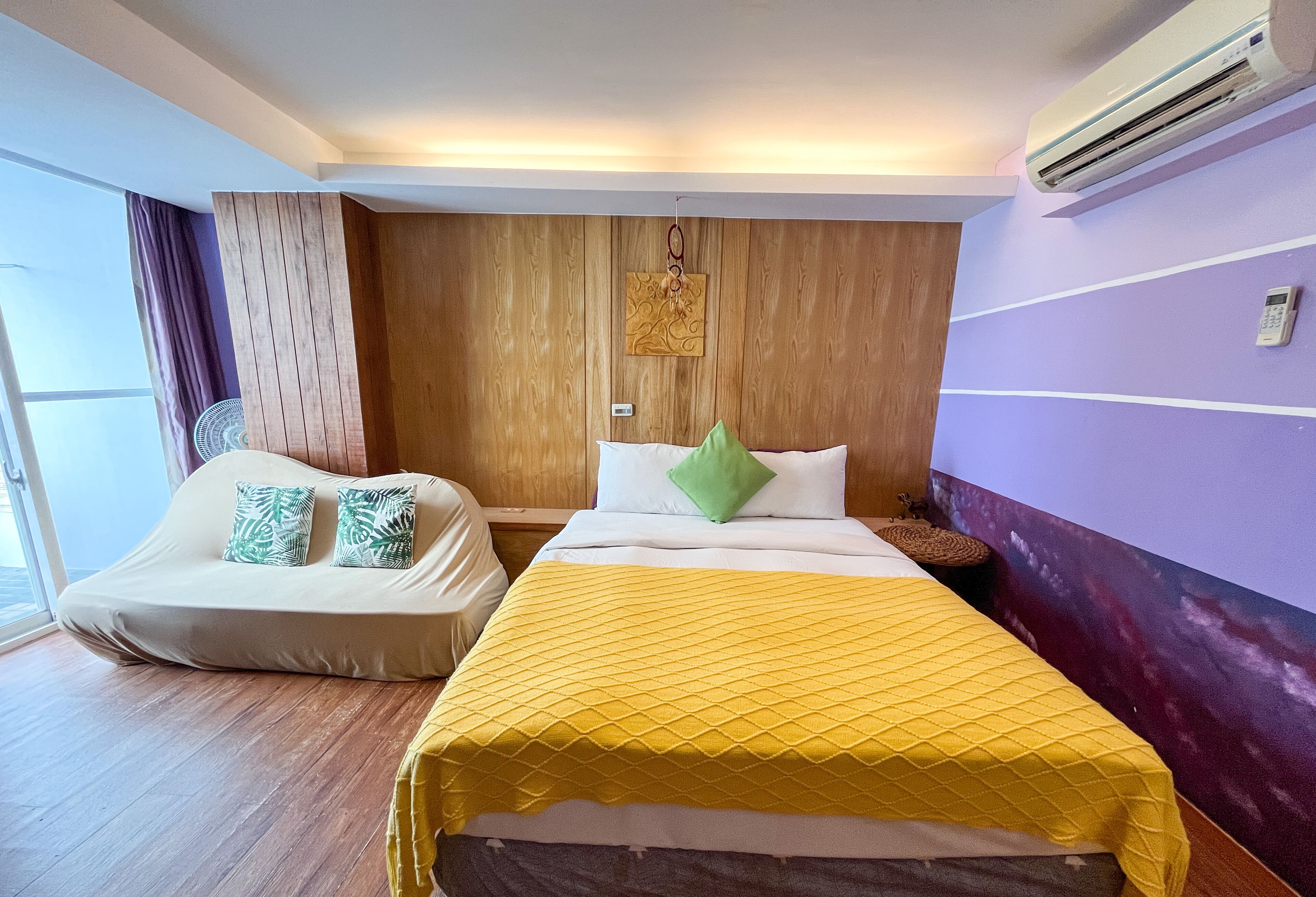 A hotel room with a bed, a sofa, a wooden panel wall, a ceiling fan, and an air conditioning unit.