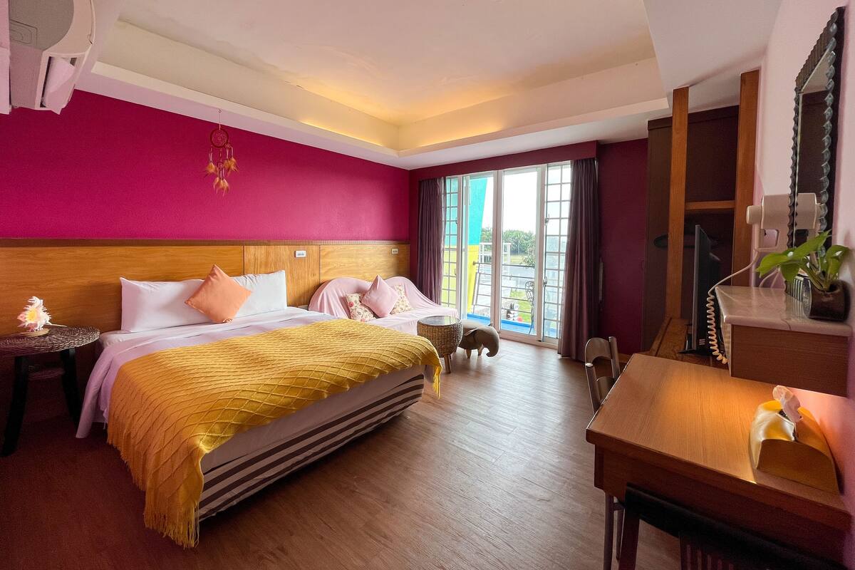 Habitación de hotel con una cama grande, cabecera de madera, mesita de noche con lámpara, escritorio con silla y una ventana amplia con cortinas.
