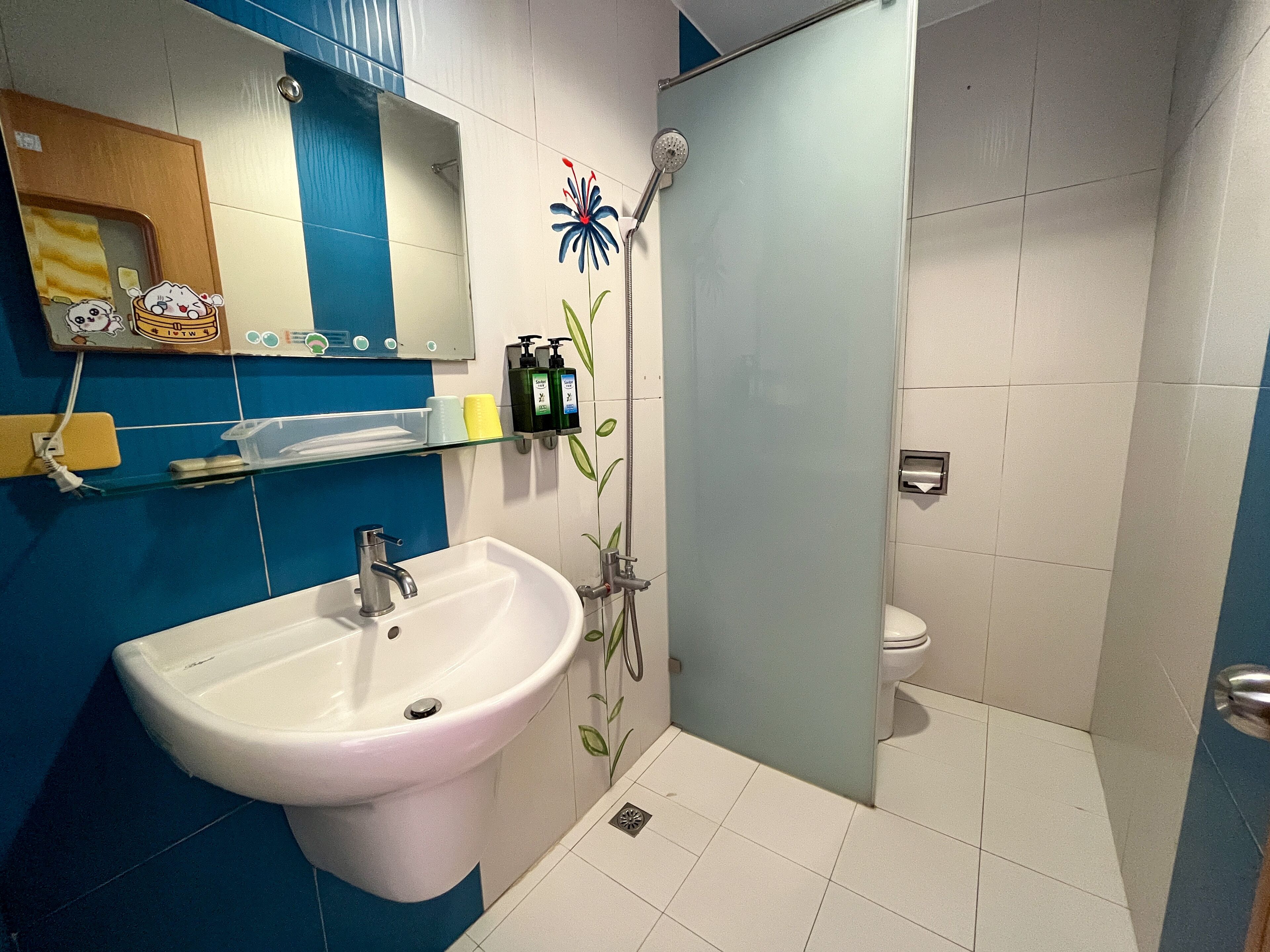 A bathroom with a sink, toilet, and shower. There are toiletries on a shelf and a mirror above the sink.