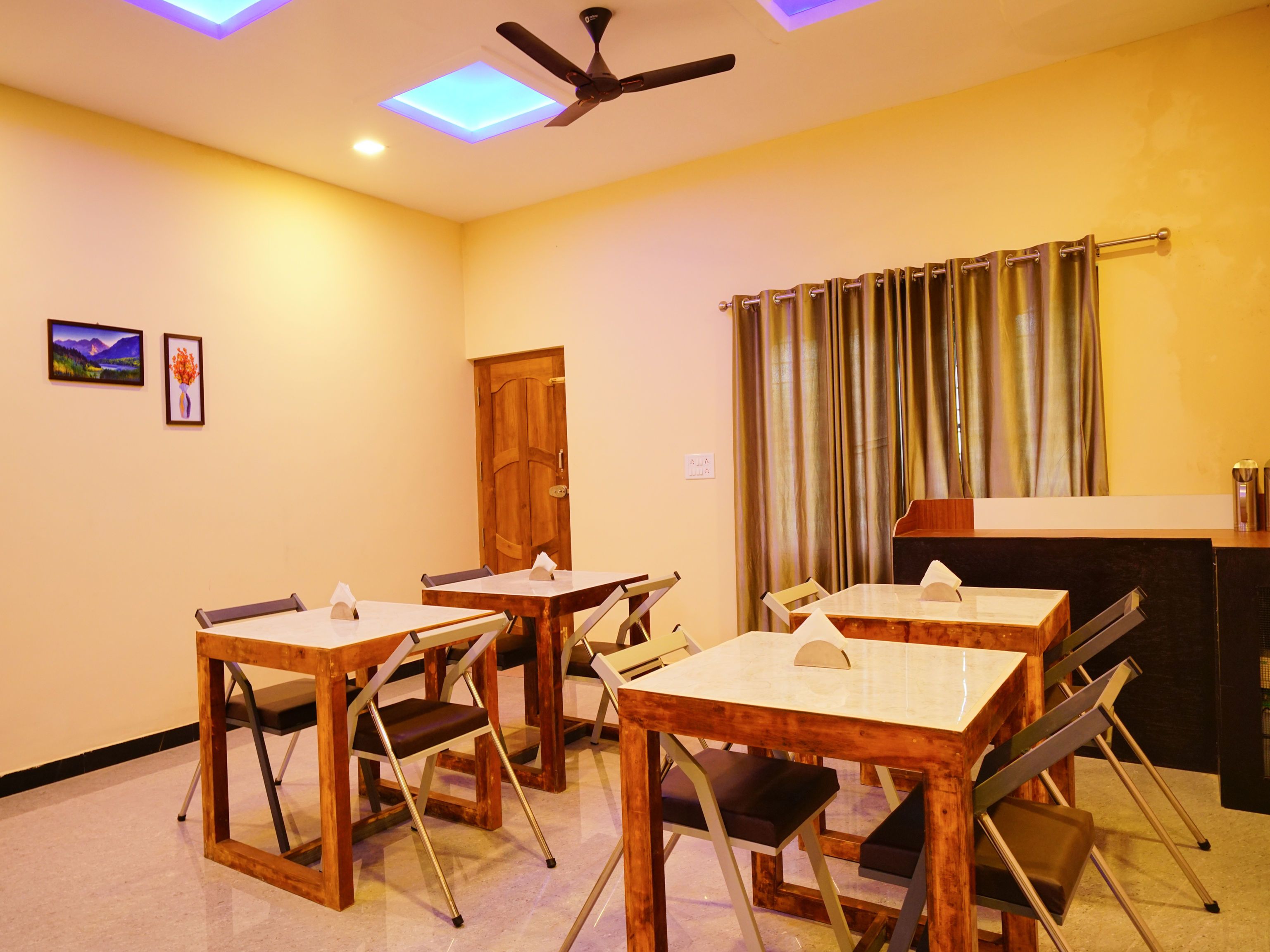A dining area with wooden tables and chairs, a ceiling fan, and a view of the outdoors through a window with curtains.