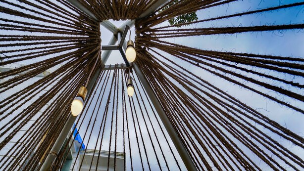 A view of a building's interior with a ceiling made of ropes, a visible light fixture, and a window showing a palm tree outside. - OYO 16630 Home Cocoa Tree Residency (Devikolam)