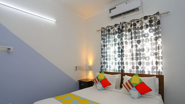 A bedroom with a bed, a nightstand, a lamp, and a window with patterned curtains. - OYO 23654 Home Cozy 2BHK Aluva Metro Station (Aluva)