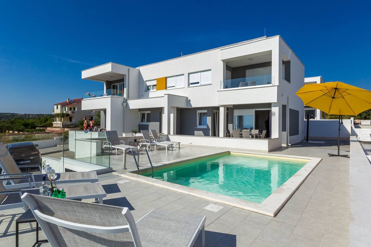 A modern two-story building with a swimming pool, outdoor seating, and a yellow umbrella.