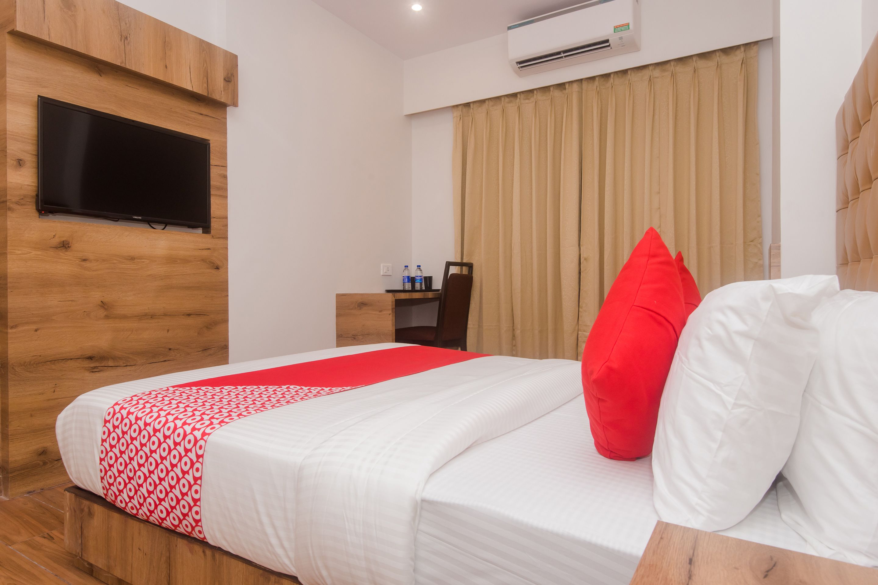 A hotel room with a bed, a TV mounted on a wooden panel, a wardrobe, and a window with curtains.