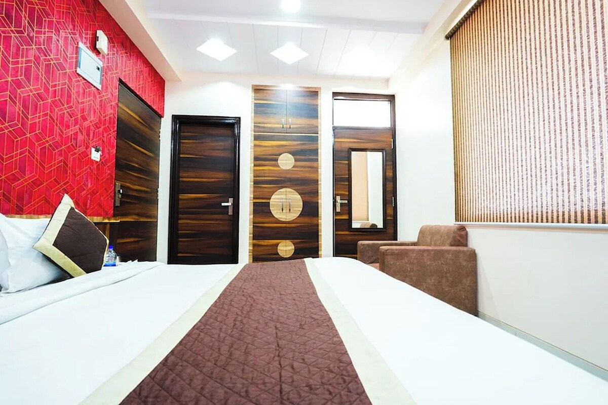A hotel room with a bed, a red patterned wall, a wooden door, a brown sofa, and a window with blinds.
