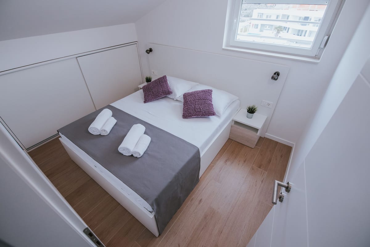 A small, modern bedroom with a bed, two pillows, a nightstand, and a window.