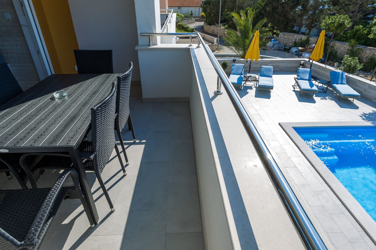 A balcony with a dining table and chairs, a pool with blue water, and yellow umbrellas.