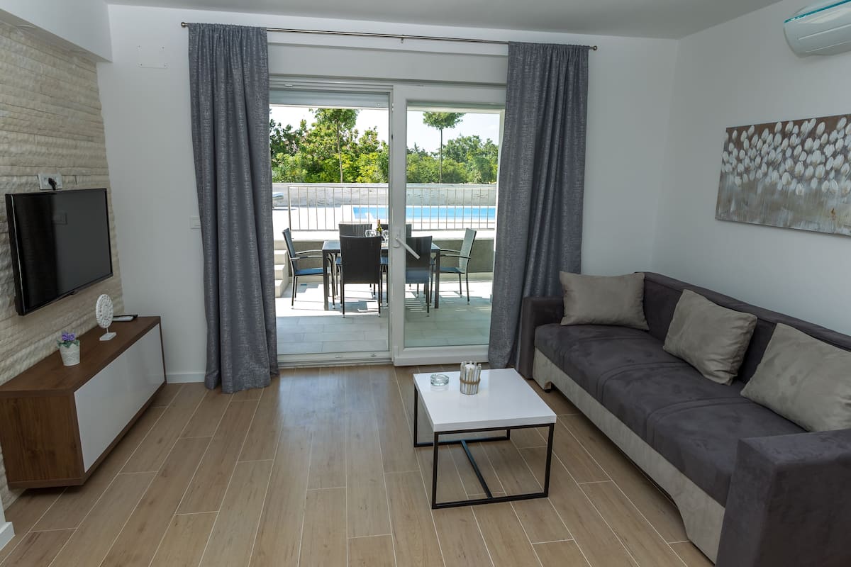 A modern living room with a flat-screen TV, a sofa, a coffee table, and a sliding glass door leading to an outdoor area with a pool.