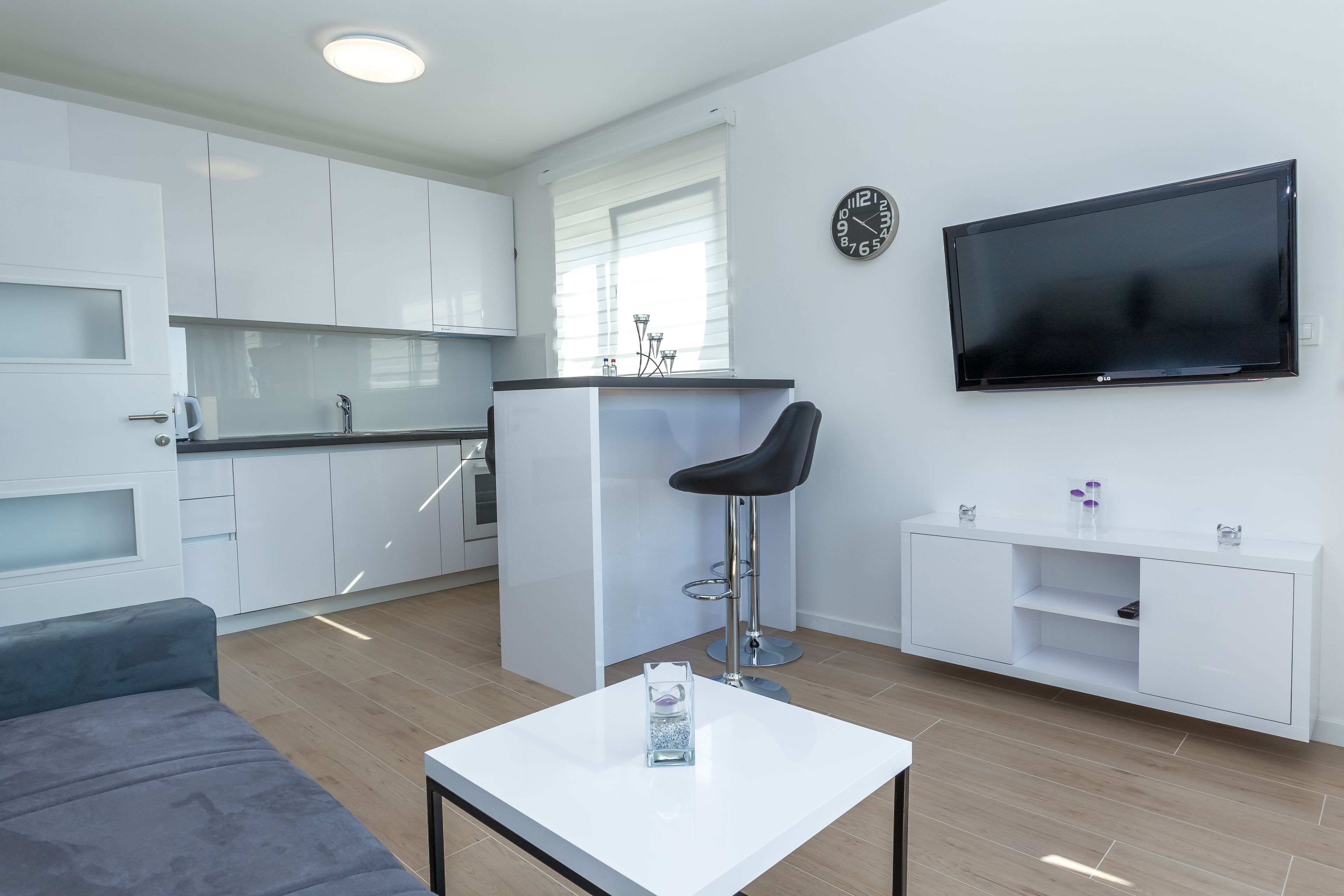 A modern kitchen with white cabinets, a black countertop, and a bar stool. A flat-screen TV is mounted on the wall, and a white console table is against the wall.