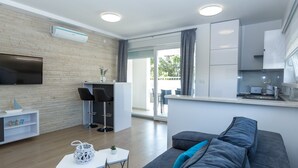 A modern living room with a grey sofa, a small white table, and a TV mounted on the wall. There is a kitchen area with white cabinets and a window. - Apartments & Rooms Villa Casia (Novalja)