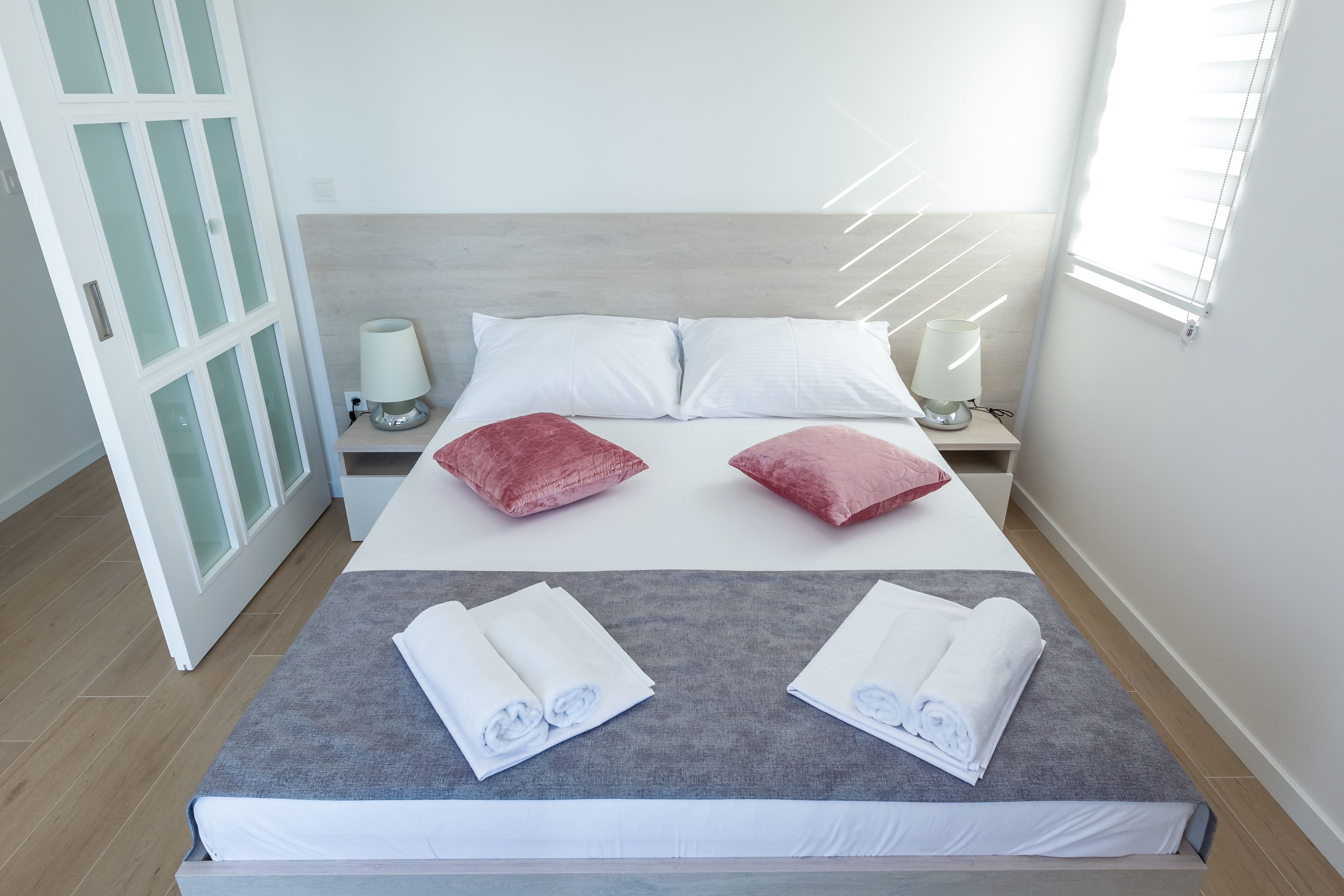 A neatly made bed with white and red pillows, two bedside lamps, and a window with blinds.