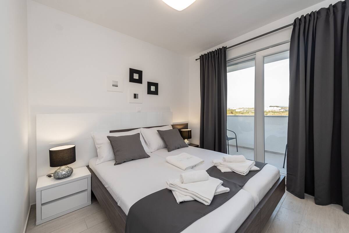 A modern hotel room with a large bed, a nightstand with a lamp, and a view of the outdoors through a large window.