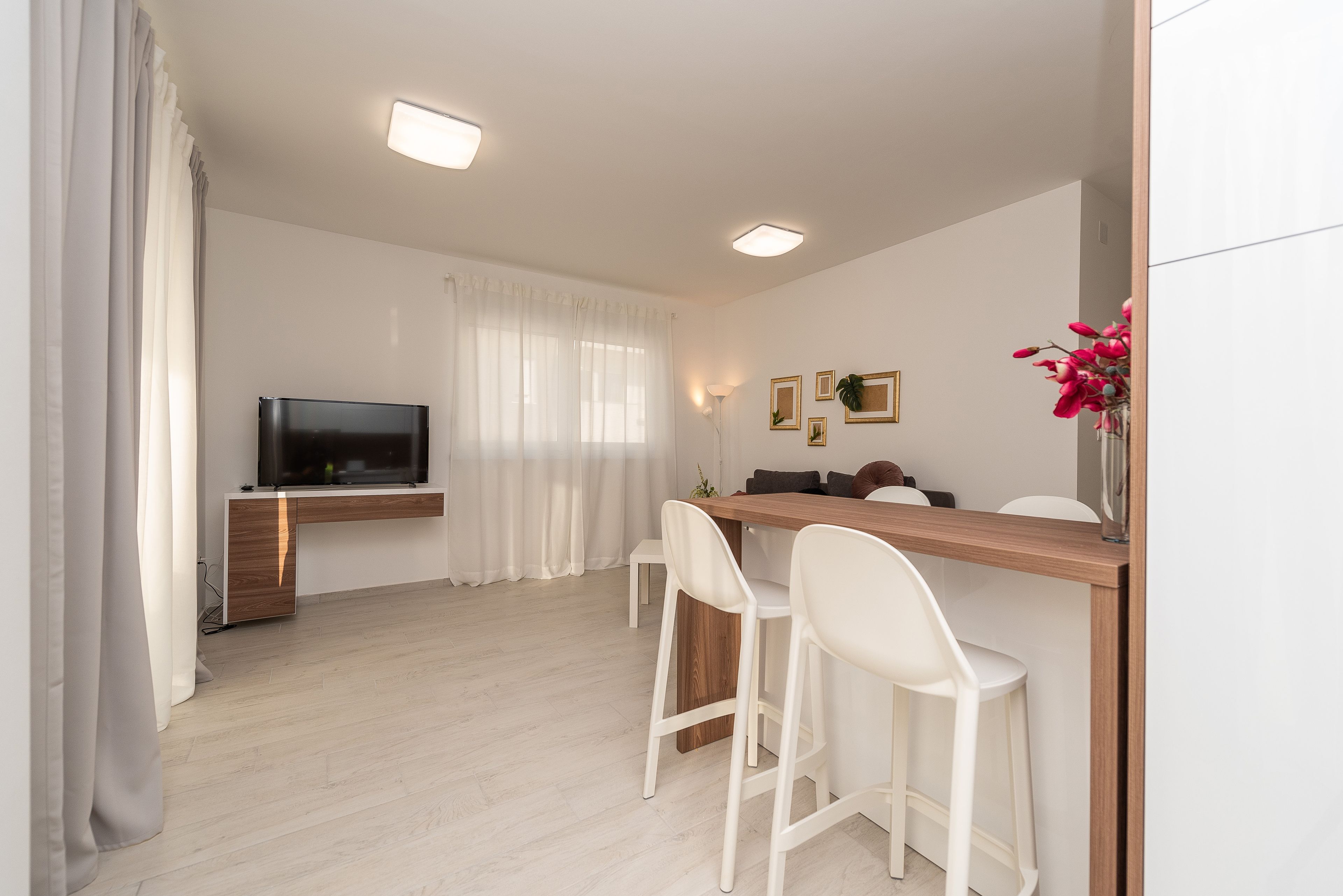 A modern living room with a flat-screen TV, a wooden dining table, white chairs, and a white sofa.