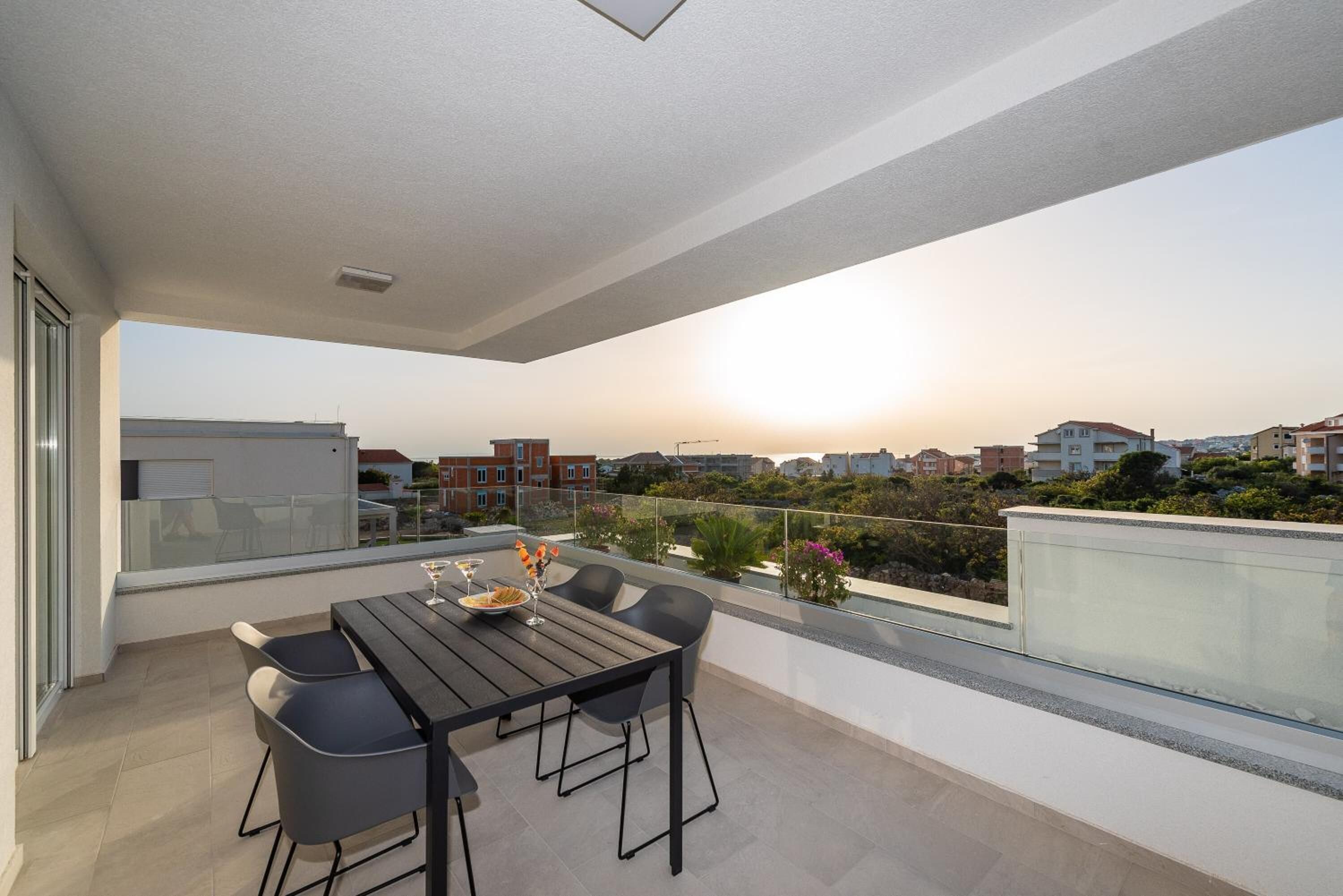 A balcony with a table and chairs, overlooking a residential area during sunset.