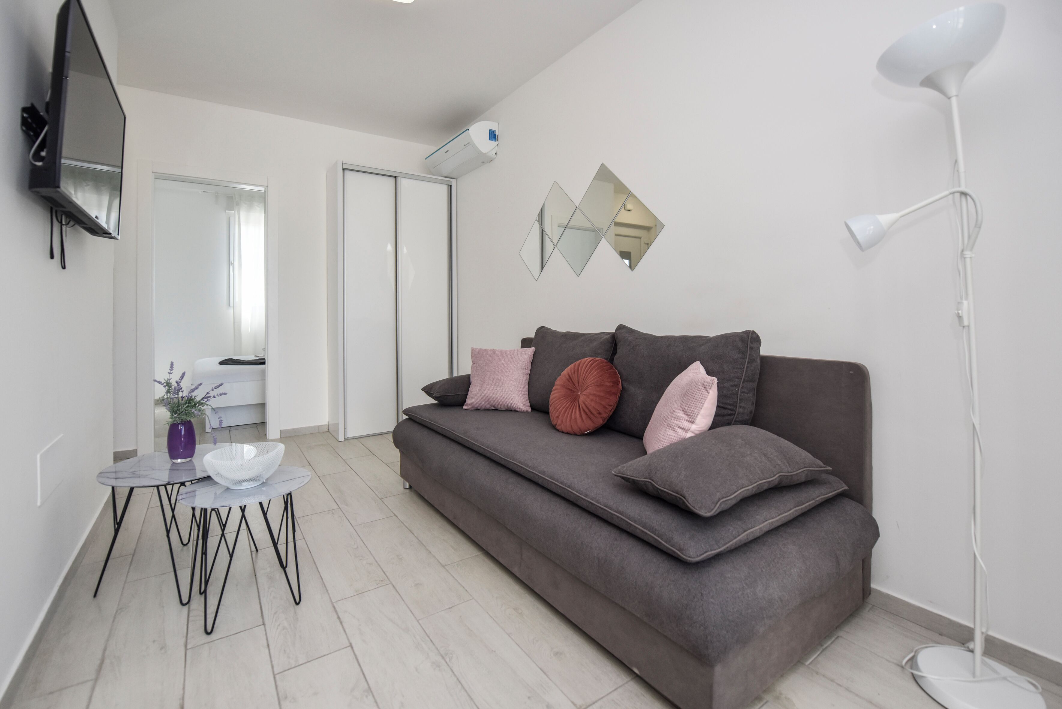 A modern living room with a grey sofa, a small round table with a glass bowl, a floor lamp, and a mounted TV.