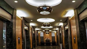 A modern hotel lobby with a marble floor, wooden paneling, and a central chandelier.