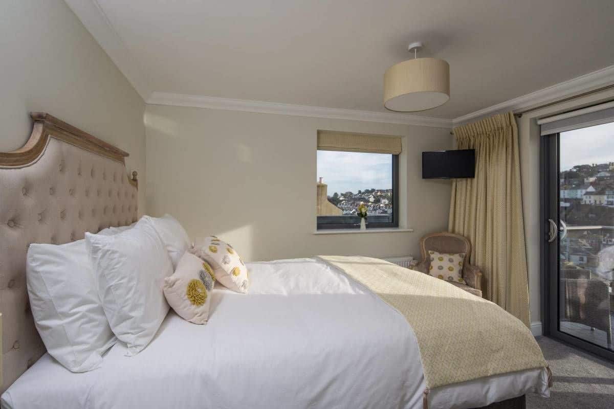 A bedroom with a large bed, a television mounted on the wall, a window with a view of buildings, and a balcony with curtains.