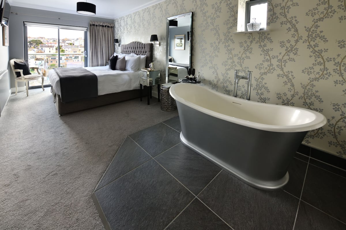 A modern bedroom with a large bed, a classic clawfoot bathtub, and a city view through a sliding door.