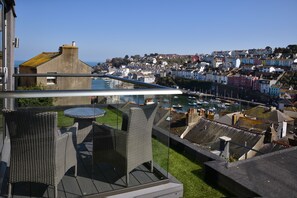 A balcony with wicker furniture and a glass barrier overlooking a coastal town with colorful houses and boats in the marina.