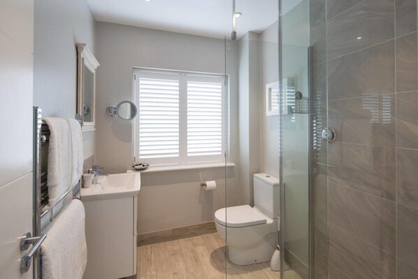 Un baño moderno con cabina de ducha de vidrio, inodoro, lavamanos con espejo y ventana con persianas.