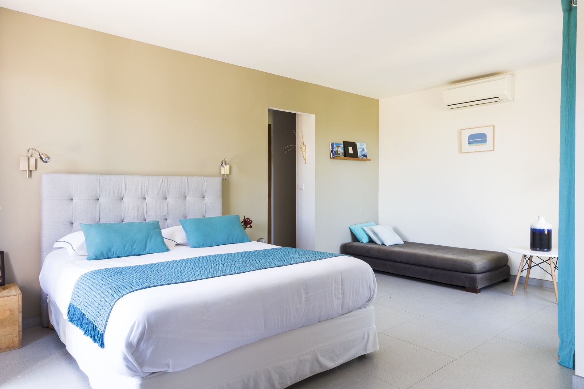 A bedroom with a large bed, a bedside table, a small table with a vase, and a grey sofa.