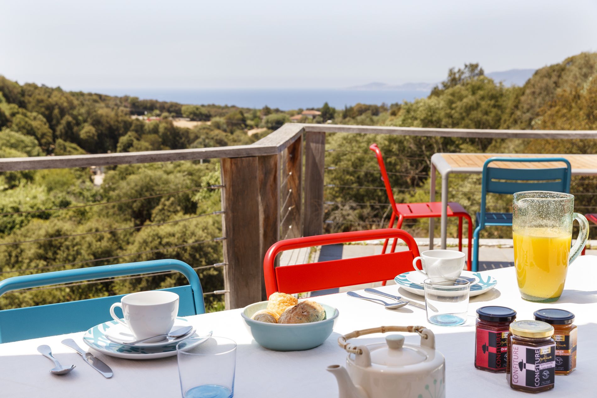 Una mesa dispuesta con alimentos para el desayuno, una tetera y condimentos, en un balcón con vistas a un paisaje boscoso.