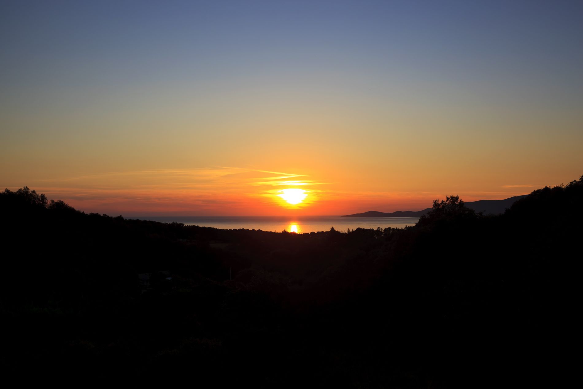 A sunset over a body of water with silhouettes of hills and trees.